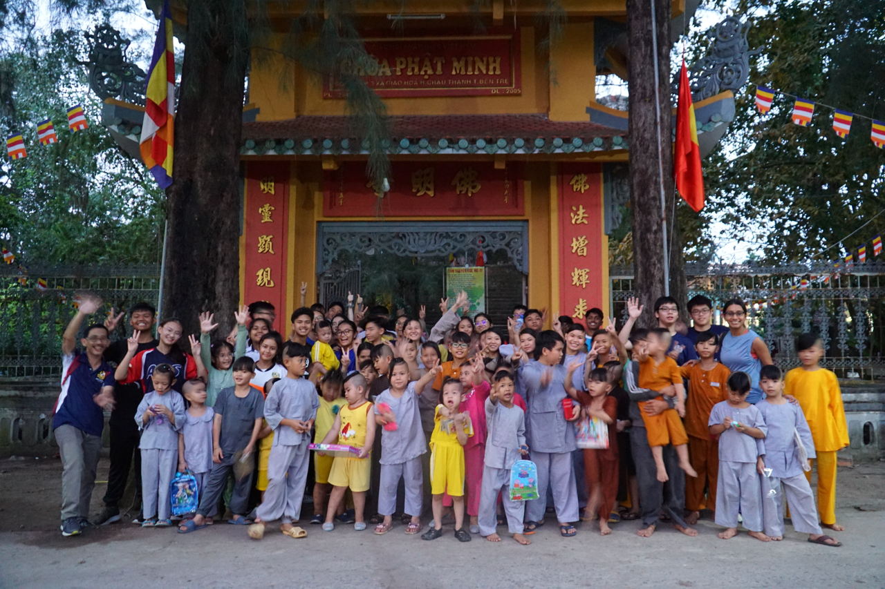 A large group of children and adults wave and pose in front of the yellow Chua Phat Minh Buddhist temple in Vietnam.